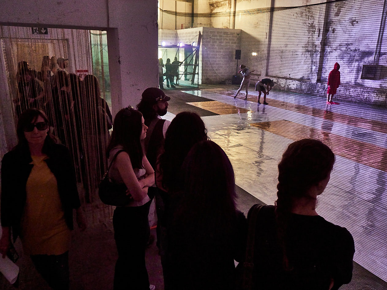 gruppo nanou – Specie di spazi. Foto: Daniele Casadio Spettatori accanto a una tenda di fili guardano i danzatori oltre una rete, una figura in rosso sul fondo