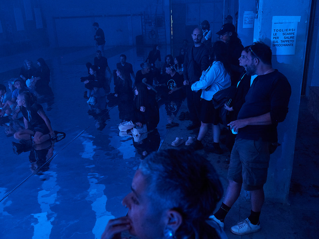 gruppo nanou – Specie di spazi. Foto: Daniele Casadio Pubblico seduto su un pavimento specchiante in luce blu, cartello che invita a togliersi le scarpe