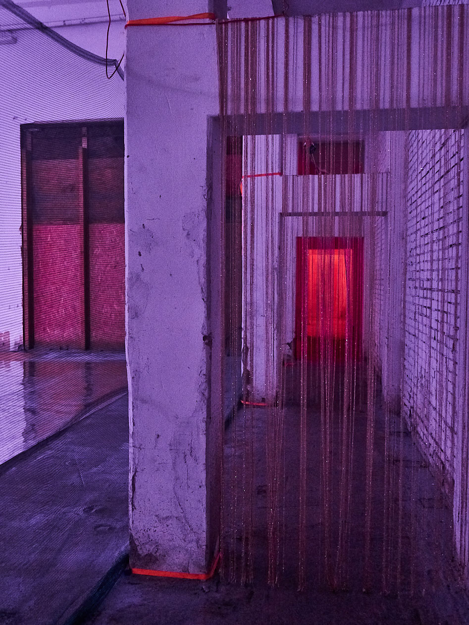 gruppo nanou – Specie di spazi. Foto: Daniele Casadio Corridoio con tende di fili in luce viola, una porta rossa illuminata in fondo