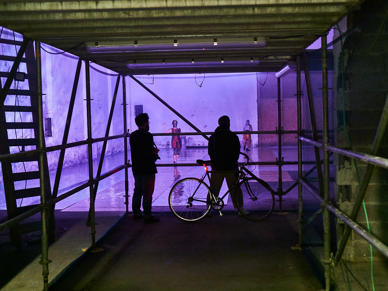gruppo nanou – Specie di spazi. Foto: Daniele Casadio Due persone e una bicicletta sotto un ponteggio, in fondo due danzatrici in luce viola