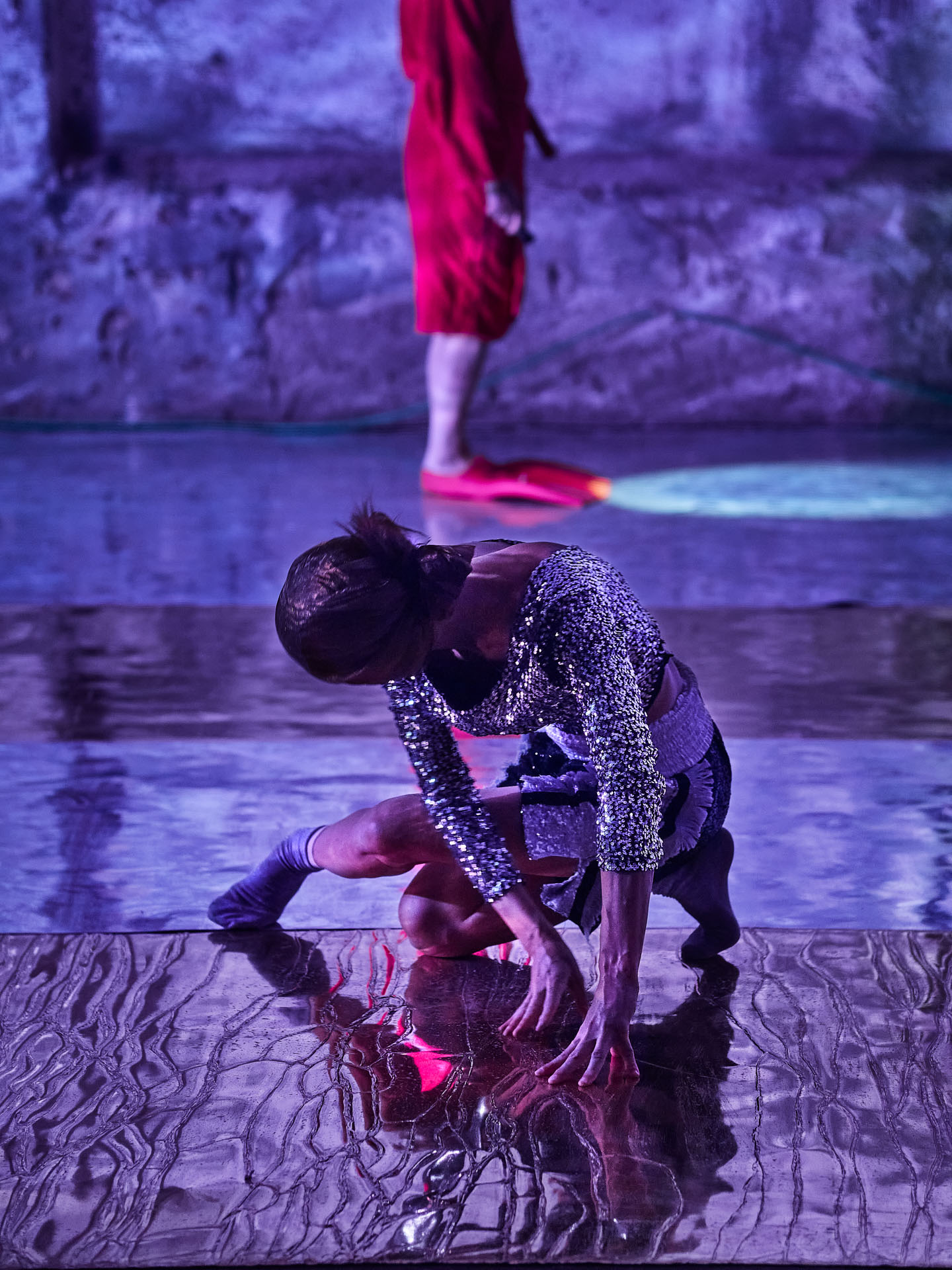 gruppo nanou – Specie di spazi. Foto: Daniele Casadio Danzatrice in paillettes accovacciata sullo specchio, alle spalle una figura in accappatoio rosso e pinne