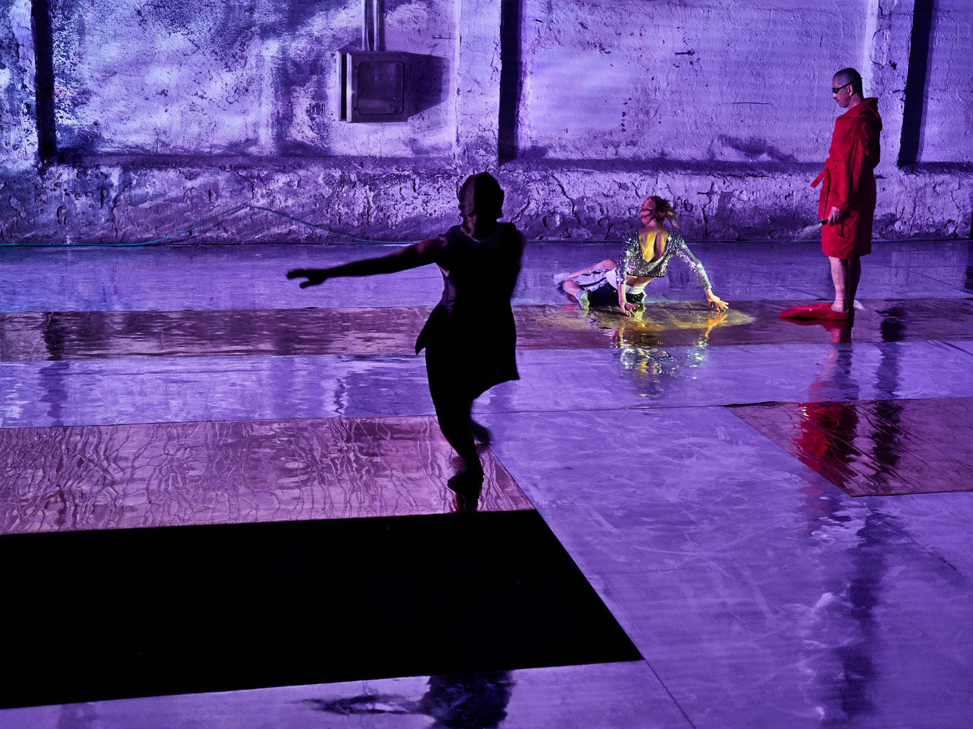 gruppo nanou – Specie di spazi. Foto: Daniele Casadio Tre performer sul pavimento specchiante in luce viola, uno in accappatoio rosso e occhiali scuri