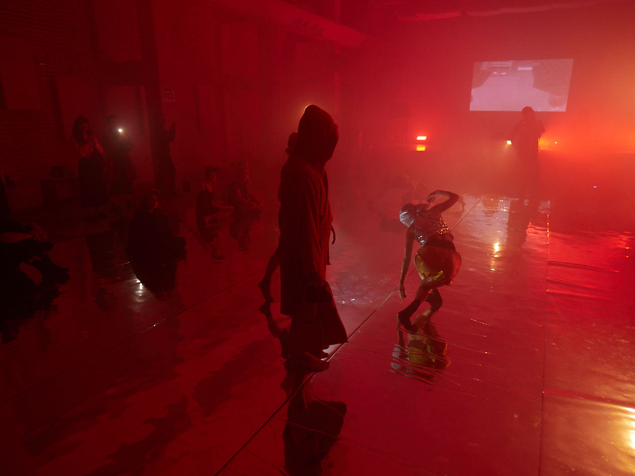 gruppo nanou – Specie di spazi. Foto: Daniele Casadio Sala immersa in luce rossa e nebbia, una figura incappucciata e una danzatrice piegata sullo specchio
